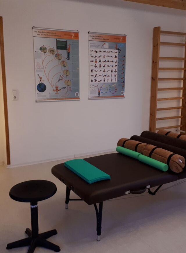 Ein Massageliege auf der Rollen in verschiedenen Größen liegen. An der Wand sind zwei Feldenkrais-Poster und eine Sprossenwand. Zwei Stehlampen beleuchten den Raum mit gedämpften Licht.
