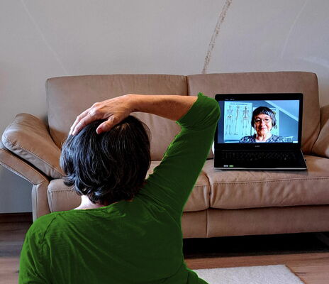 Eine Frau macht eine Feldenkrais-Übung. Sie sitzt auf dem Boden mit dem Rücken zur Kamera. Ihre rechte Hand ist auf dem Kopf und sie schaut auf einen Laptop, der vor ihr auf einem Ledersofa steht.